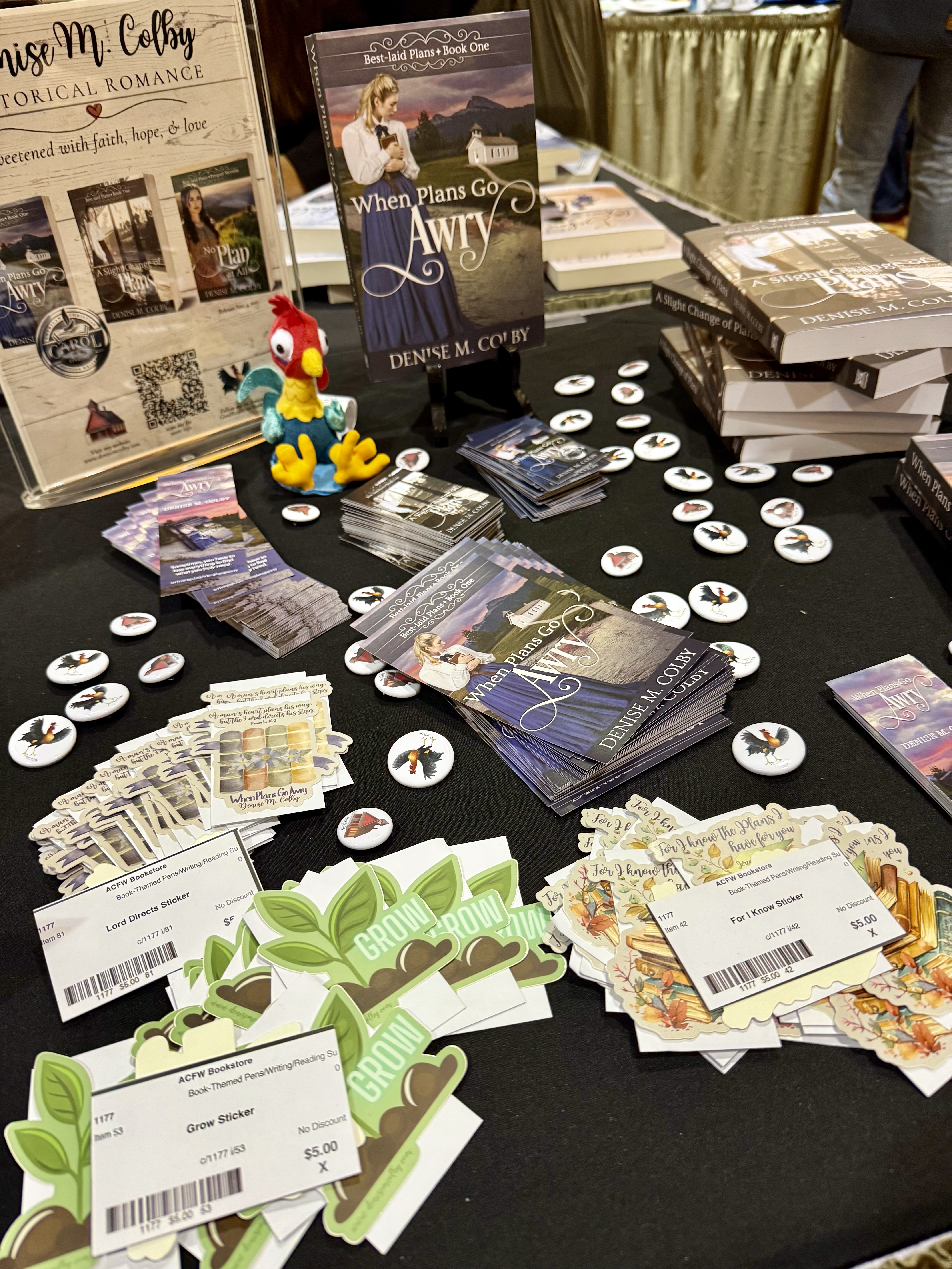 Denise M. Colby tabletop display including stickers, books, bookmarks, business cards, and Hei Wei, the rooster from Moana to represent BERT in her stories